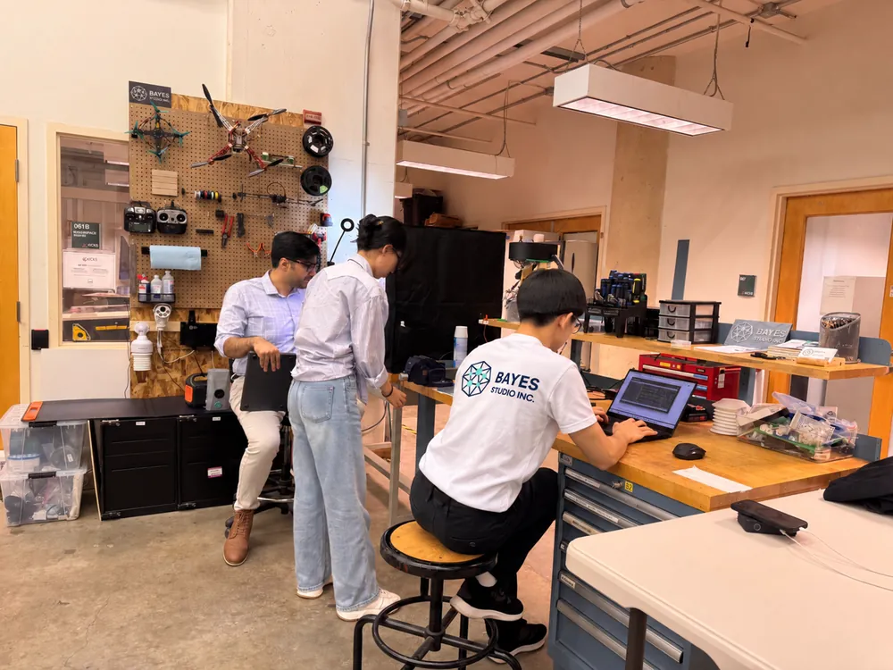 Three of the Bayes Studio team working in the UBC HATCH workshop: two standing at a bench under a pegboard hung with drone airframes and tools, one seated at a laptop in a Bayes Studio Inc. shirt.